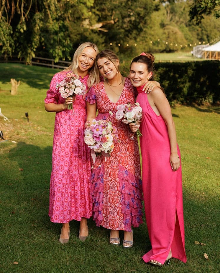 Three bridesmaids in elegant pink dresses holding blush floral bouquets, smiling together during an English countryside wedding reception
