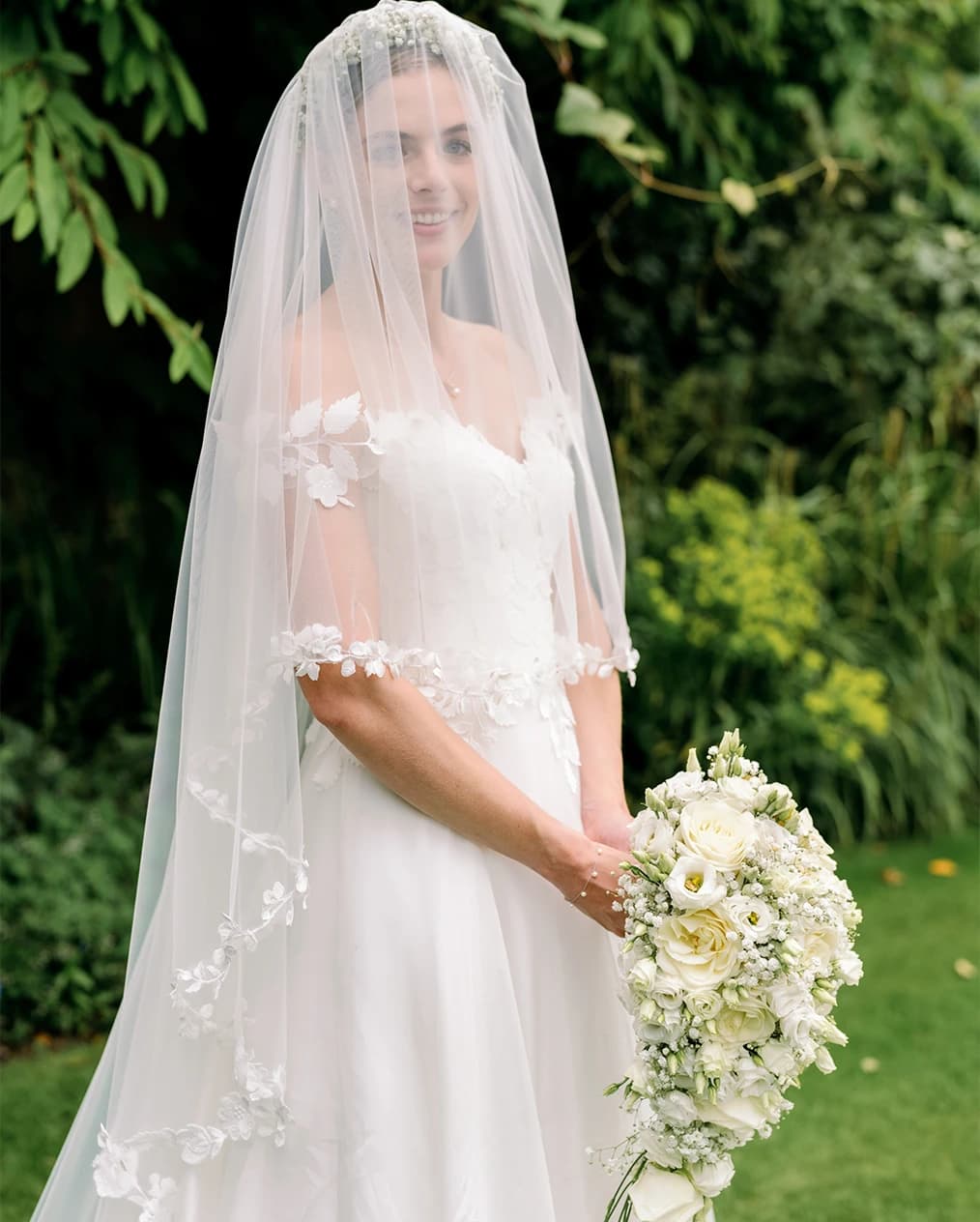 Bride wearing dress and veil with 3D florals, while holding a bouquet of white roses and gypsophila