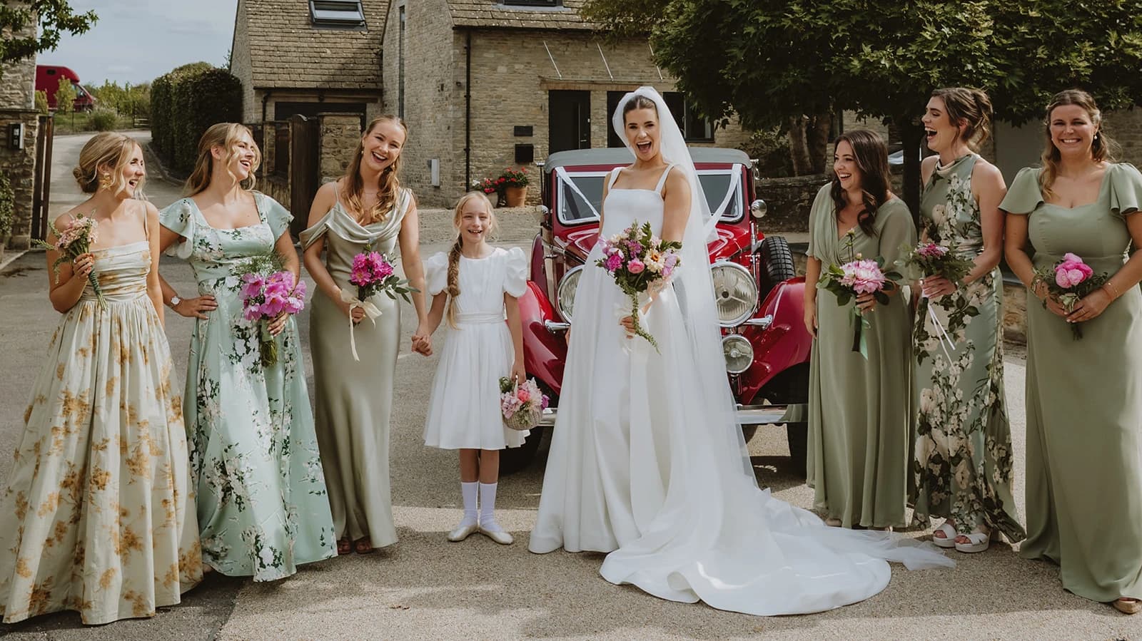 Bride, bridesmaids and flower girl gathered beside the red Alvis at St Edburg's Church