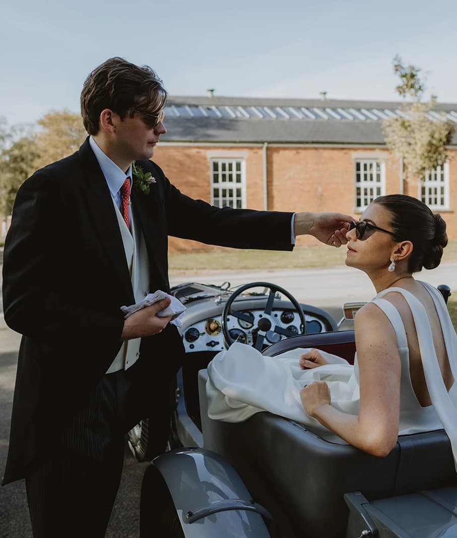Groom straightening the bride's sunglasses as she sits in a vintage Bentley
