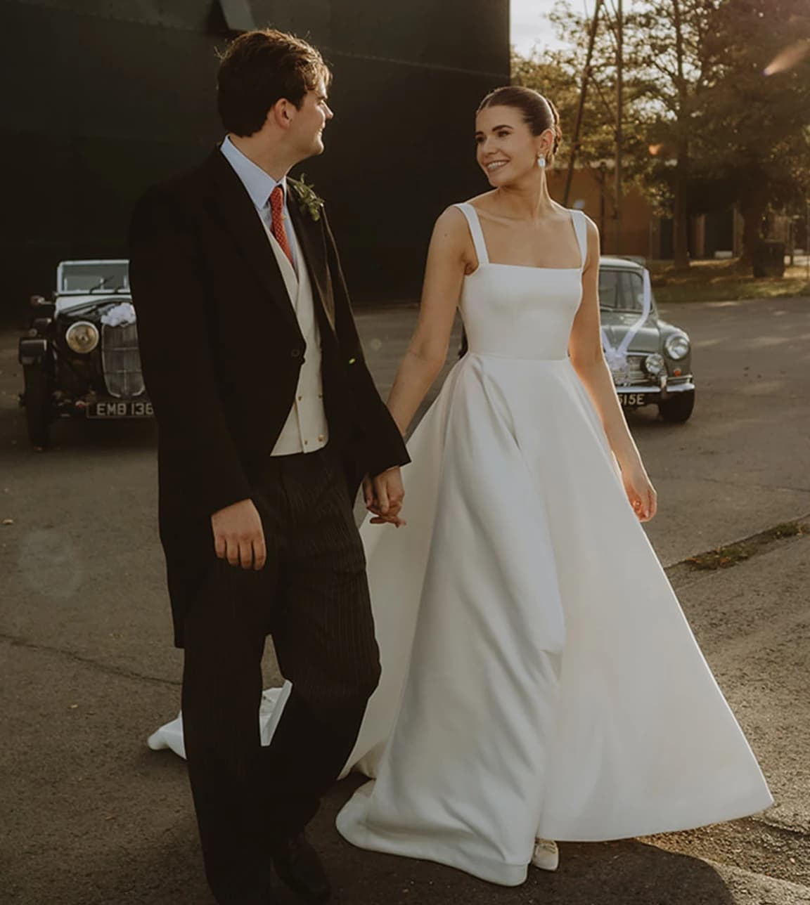Bride in a square-neck wedding dress walking hand in hand with the groom past classic cars at Bicester Motion in golden light