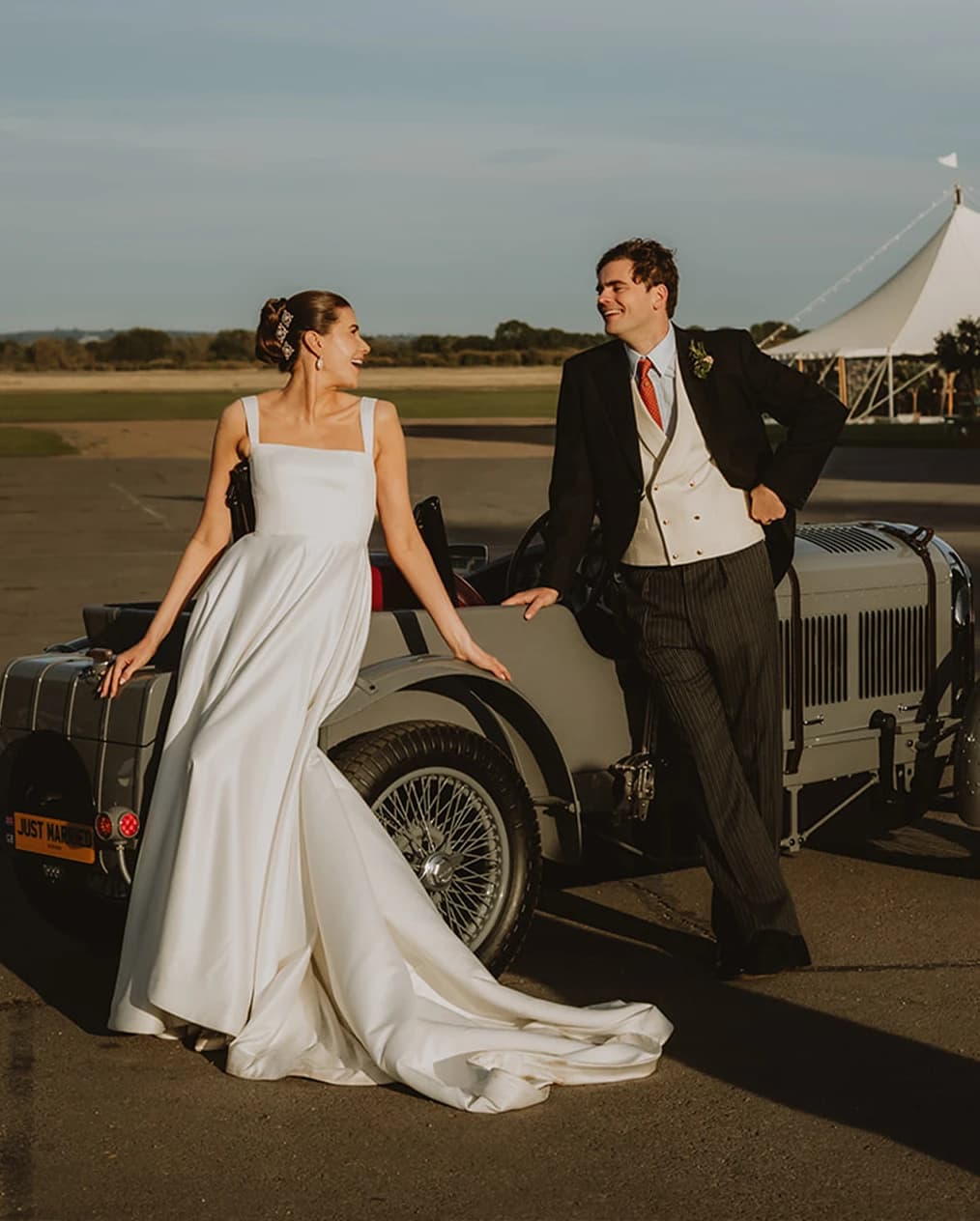 Bride and groom laughing as they lean on a grey vintage car with a Just Married plate, the marquee behind on the Bicester Motion airfield